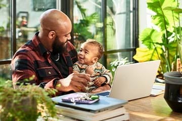 woman holds child while infront of computer and looking at phone with other children in background