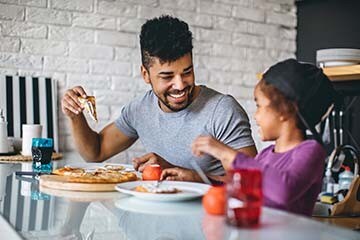 A father and daughter are smiling while eating pizza together in their kitchen.