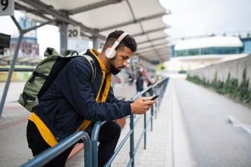 A young man is wearing headphones and looking at his phone while at a bus station.