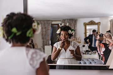A bride is looking in a mirror and putting on lipstick. In the background, a photographer snaps a picture.