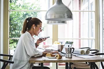 Woman with a ponytail sitting at her kitchen table on her laptop and taking a call with her wireless headphones.