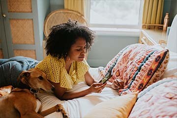 Image of a woman looking at her phone while lounging on her bed with her dog