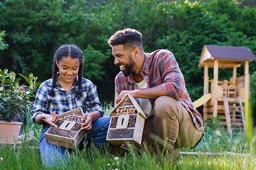 A young man is in a backyard with his younger sister. They are each holding bug hotels and smiling.