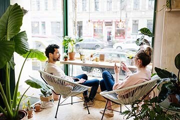 A couple is sitting together inside a café decorated with plants. They are next to a large window and it is sunny outside.