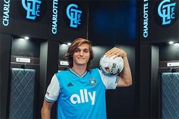  Charlotte FC midfielder, Ben Bender, poses in front of Charlotte FC logos in the team locker room while holding a soccer ball by his shoulder