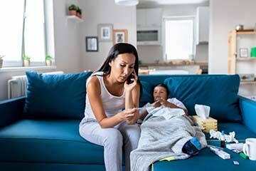 Woman holding a thermometer while on the phone and a sick child next to her.