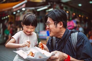 A father and daughter are smiling while eating street food on vacation. 