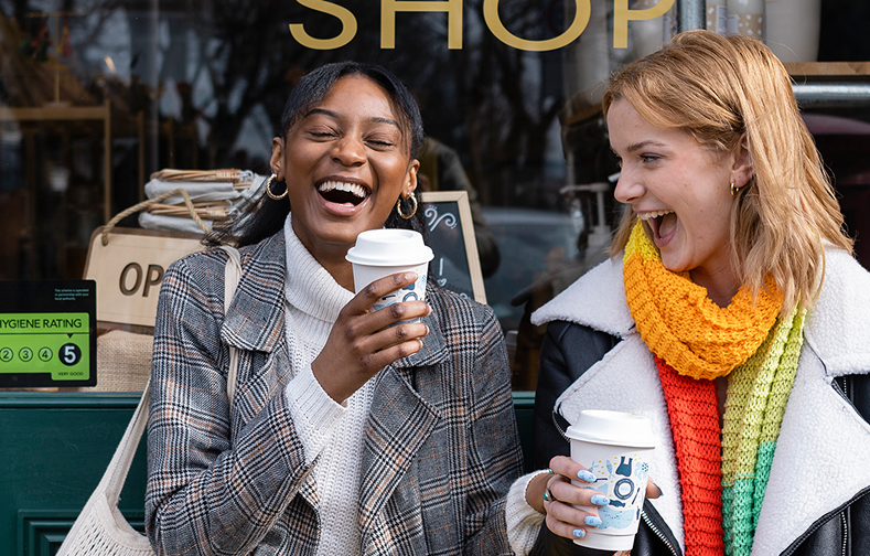 Two friends sitting in front of a shop laughing while holding coffee.