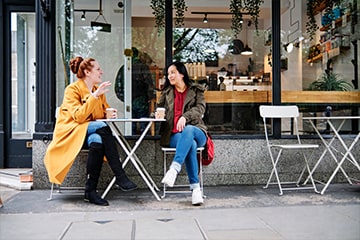 Two women drink coffee and chat outside of a cafe